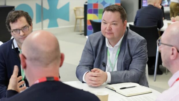 A group of three people is engaged in a discussion at a round table. One person, wearing a grey blazer, appears to be listening attentively while another two are facing him, one with short hair and the other bald. They are in a brightly lit, modern environment with colourful decorations in the background. One person has a notebook and a pen on the table.