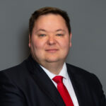 A studio portrait of a man wearing a dark suit jacket, white shirt, and bright red tie. He faces the camera with a neutral, slight smile and short dark hair neatly styled. The background is plain grey, and the lighting is even and professional, creating a formal headshot-style image